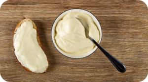 Slice of bread with mayo spread next to a bowl of mayo with spoon in it